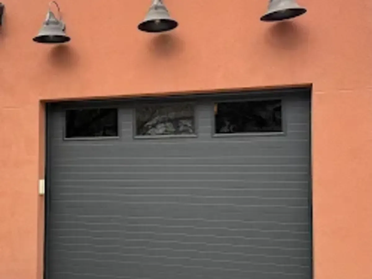 A charcoal flush-panel garage door with three top windows set into an orange stucco wall